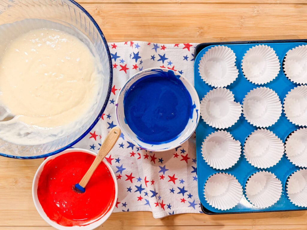 Bowl of white cake batter, bowls of red and blue batter with spatulas, and a tray with white cupcake liners on a star-patterned cloth—perfect for making festive Red White and Blue Cupcakes on a wooden surface.