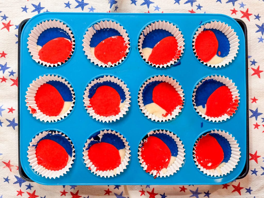 A blue muffin tin holds twelve cupcake liners filled with vibrant batter for Red White and Blue Cupcakes, sitting on a cloth decorated with red and blue stars.
