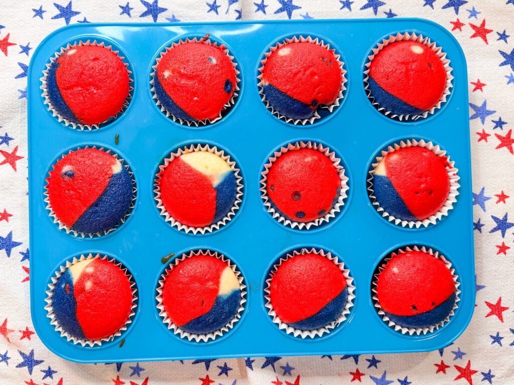 A blue muffin tin holds twelve Red White and Blue Cupcakes. The cupcakes are mostly red on top with blue and some white swirled sections, resting on a star-patterned cloth.