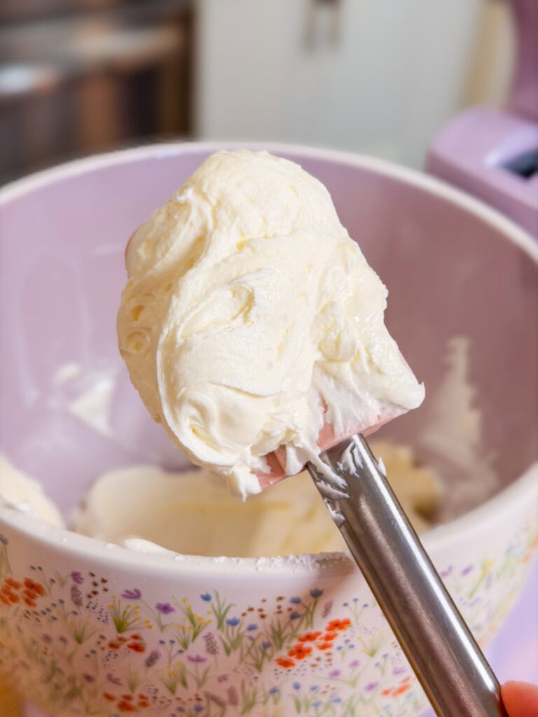 A metal spatula holds a dollop of creamy white frosting above a pastel mixing bowl decorated with colorful floral patterns, perfect for topping festive Red White and Blue Cupcakes.