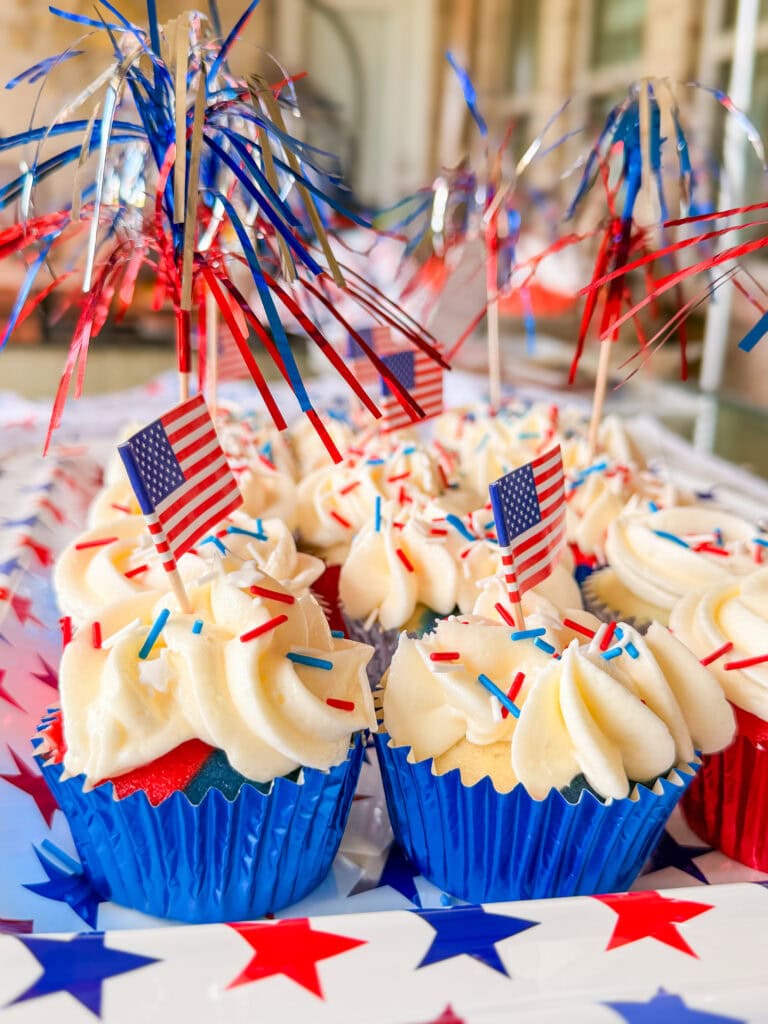 Red White and Blue Cupcakes with white frosting, red and blue sprinkles, and small American flags in blue foil wrappers, displayed on a table decorated with stars and festive tinsel toppers for a patriotic touch.