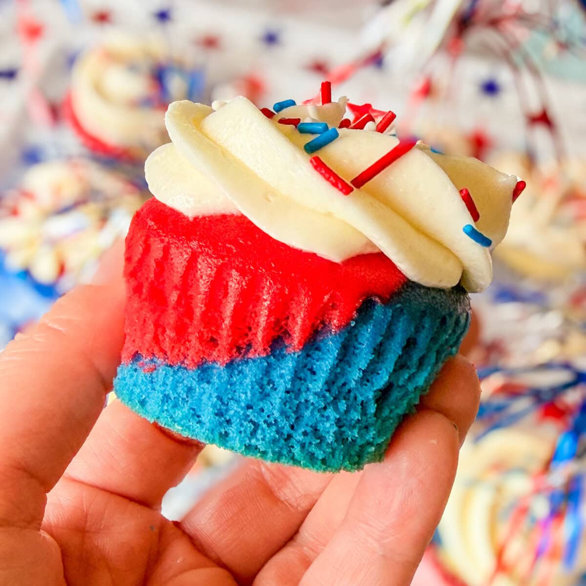 A hand holds a Red White and Blue Cupcake with blue and red layers, topped with white frosting and festive sprinkles. The background features more Red White and Blue Cupcakes and cheerful star patterns.