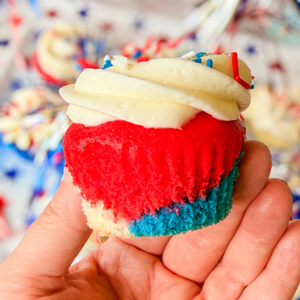 A hand holds one of the Red White and Blue Cupcakes with white frosting and sprinkles. The colorful layers are visible from a bite taken out, while festive decorations blur in the background.