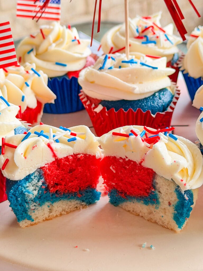 Close-up of Red White and Blue Cupcakes with patriotic cake layers, topped with white frosting and colorful sprinkles. Mini American flags on toothpicks decorate some cupcakes, and one is cut in half to reveal the vibrant tri-color inside.