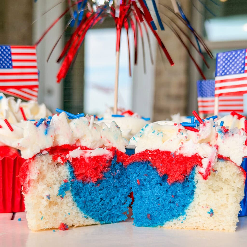 A Red White and Blue Cupcake is cut in half, showing a blue and white swirl inside. The cupcake features festive frosting, while small American flags and patriotic decorations add flair in the background.