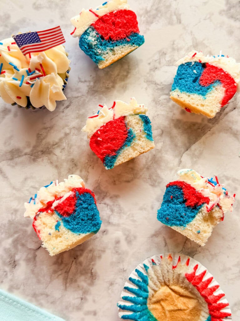 Red White and Blue Cupcakes, each cut in half to reveal their vibrant marbled interior, are topped with white frosting and patriotic sprinkles. One cupcake features a small American flag toothpick, with a used wrapper nearby.