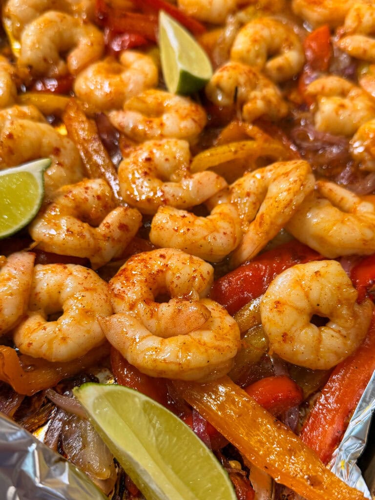 Close-up of Sheet Pan Shrimp Fajitas with seasoned shrimp and sautéed bell peppers on a foil-lined tray, garnished with lime wedges. The dish features vibrant colors and a glossy, freshly-cooked appearance.