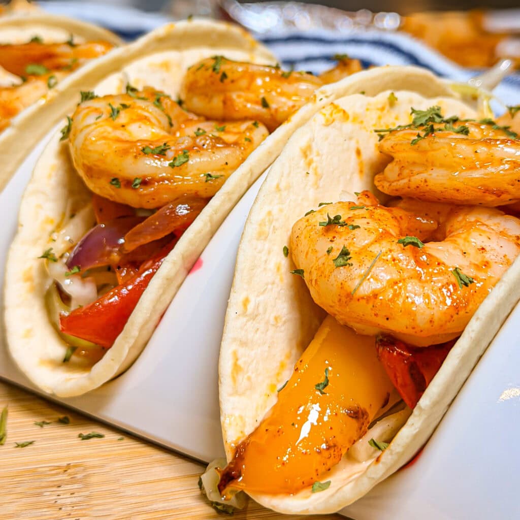 Close-up of Sheet Pan Shrimp Fajitas with sautéed bell peppers and onions in soft flour tortillas, garnished with chopped herbs, served on a white taco holder atop a wooden surface.