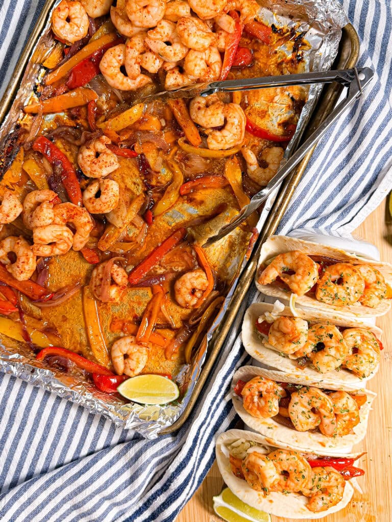 A sheet pan with Sheet Pan Shrimp Fajitas—cooked shrimp, bell pepper strips, and onions—next to three shrimp tacos in tortillas, garnished with herbs, on a striped cloth with lime wedges nearby.