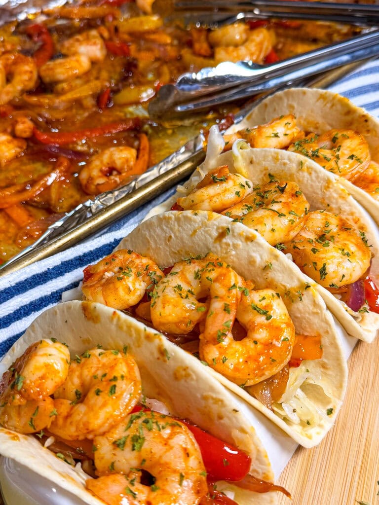 Three shrimp tacos in flour tortillas, garnished with herbs and filled with shrimp and sautéed vegetables, sit on a wooden board—bringing the flavors of Sheet Pan Shrimp Fajitas to your table—with a tray of cooked shrimp and peppers in the background.