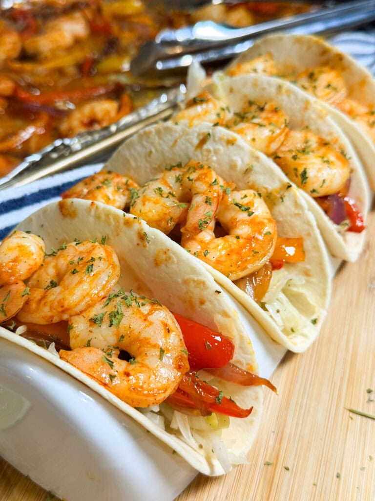 Three shrimp tacos in soft tortillas, filled with cooked shrimp, sliced red peppers, and shredded cabbage, garnished with herbs, evoke the flavors of Sheet Pan Shrimp Fajitas. Tongs and more shrimp are visible in the background.