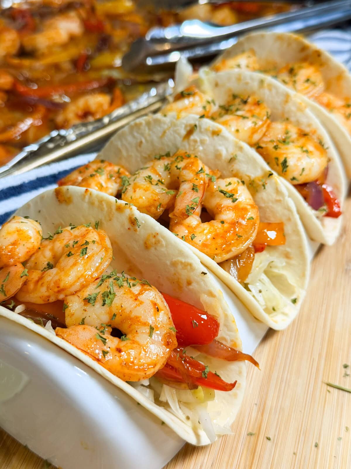 Three shrimp tacos in soft tortillas, filled with cooked shrimp, sliced red peppers, and shredded cabbage, garnished with herbs, evoke the flavors of Sheet Pan Shrimp Fajitas. Tongs and more shrimp are visible in the background.
