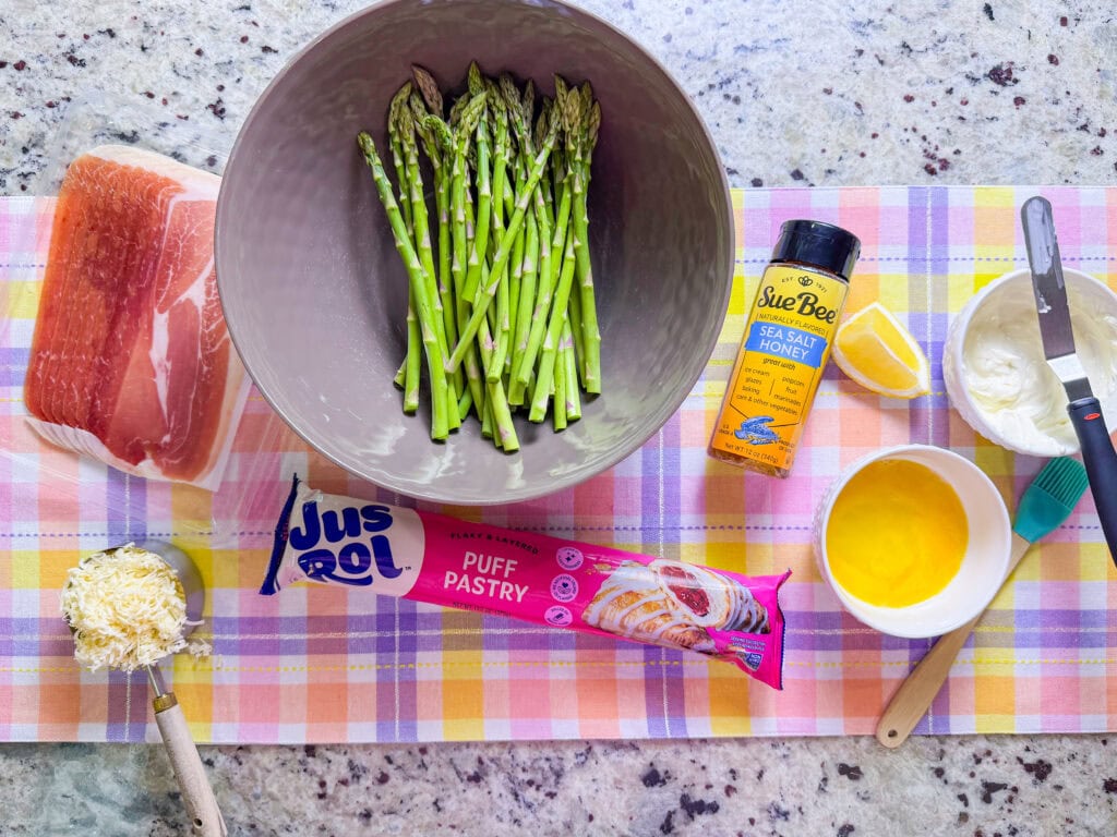 Top-down view of ingredients for a Puff Pastry Asparagus Tart, including asparagus in a bowl, prosciutto, shredded cheese, lemon, honey, sea salt, and crème fraîche—all artfully arranged on a colorful plaid cloth.