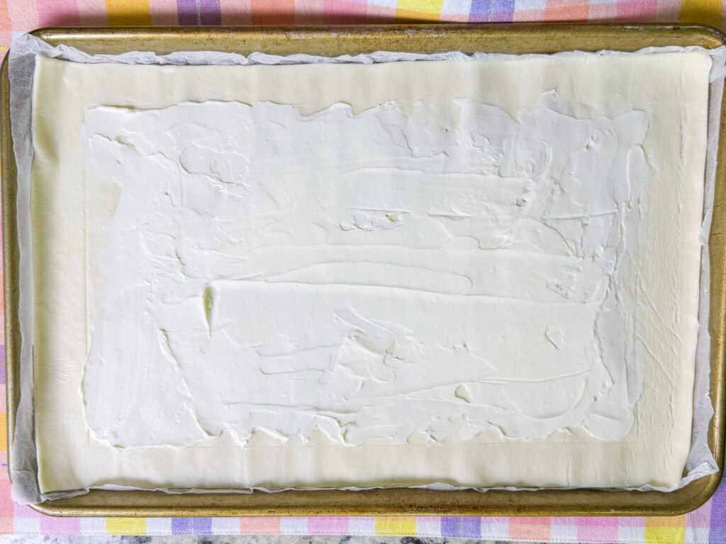 A baking sheet lined with parchment paper holds a rectangular Puff Pastry Asparagus Tart, evenly spread with a layer of white cream cheese mixture. The setup rests on a colorful, checkered cloth.