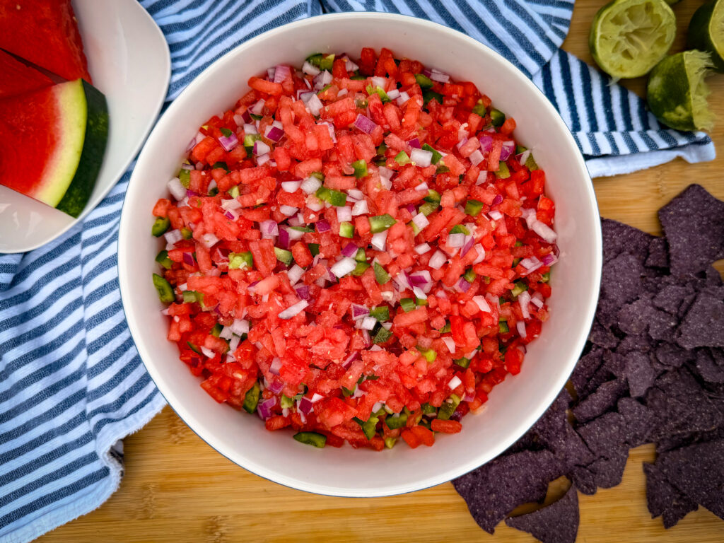 A bowl of The Best Watermelon Salsa with diced watermelon, red onion, and jalapeño sits on a striped cloth, surrounded by blue corn chips, watermelon slices, and squeezed limes on a wooden surface.
