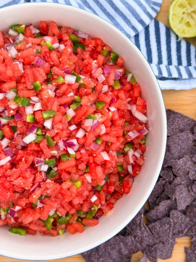 A bowl of The Best Watermelon Salsa, made with diced watermelon, red onion, and green pepper, sits on a wooden surface next to blue corn tortilla chips, a halved lime, and a blue-and-white striped towel.
