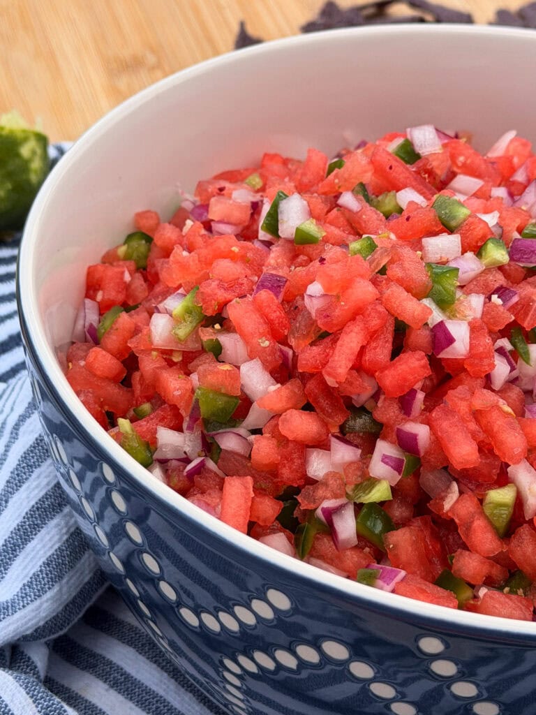 A close-up of a blue and white patterned bowl filled with The Best Watermelon Salsa—featuring diced watermelon, red onion, and jalapeño—set on a blue-striped cloth.