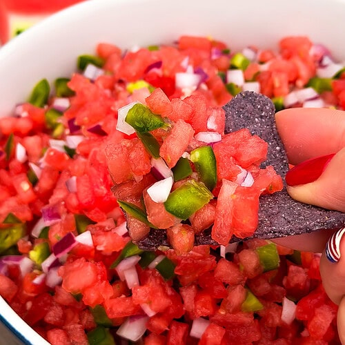 A hand with red-painted nails holds a blue tortilla chip topped with diced watermelon, onions, and jalapeños from a bowl of the best watermelon salsa.
