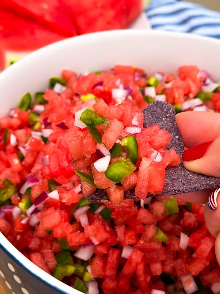 A hand with red-painted nails holds a blue tortilla chip topped with diced watermelon, onions, and jalapeños from a bowl of the best watermelon salsa.