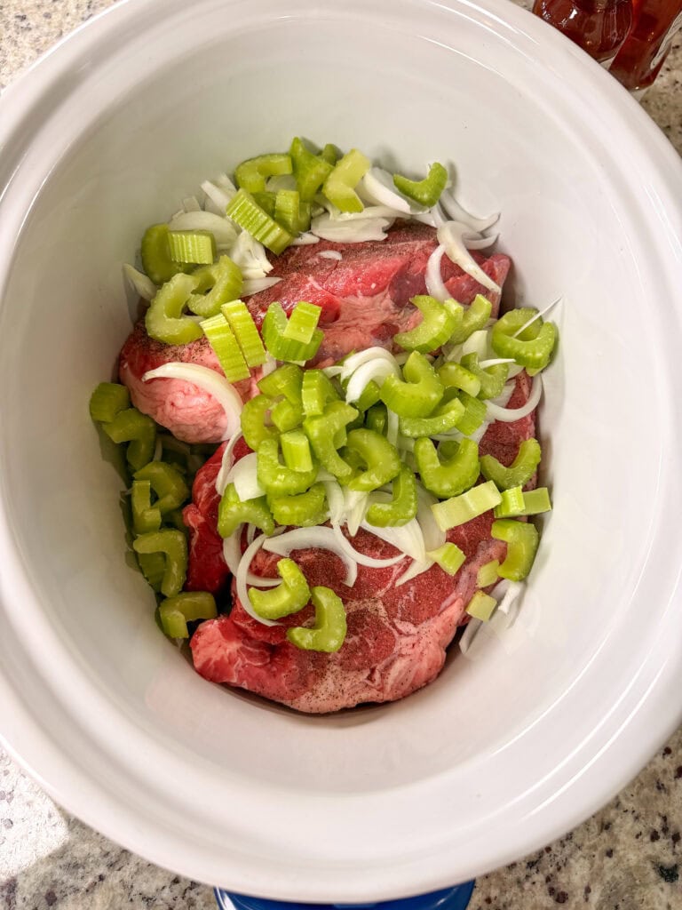 Two raw beef roasts in a white slow cooker, prepped for Crock Pot Italian Beef Sandwiches, are topped with chopped celery and sliced onions, sitting on a speckled countertop.