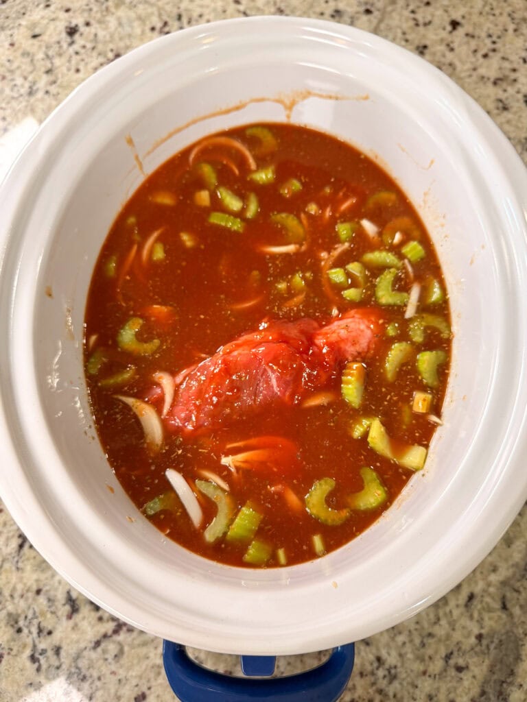 A Crock Pot Italian Beef Sandwiches recipe awaits, with a slow cooker filled with red tomato-based sauce, raw pork roast, chopped celery, and sliced onions sitting on a speckled granite countertop.