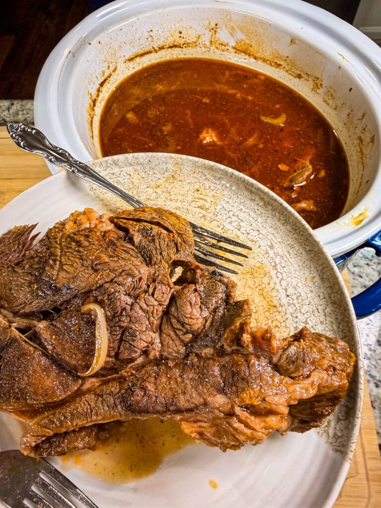 A large piece of cooked beef on a plate with two forks, in front of a slow cooker filled with red broth and onions—perfectly tender and ready to be shredded for Crock Pot Italian Beef Sandwiches.
