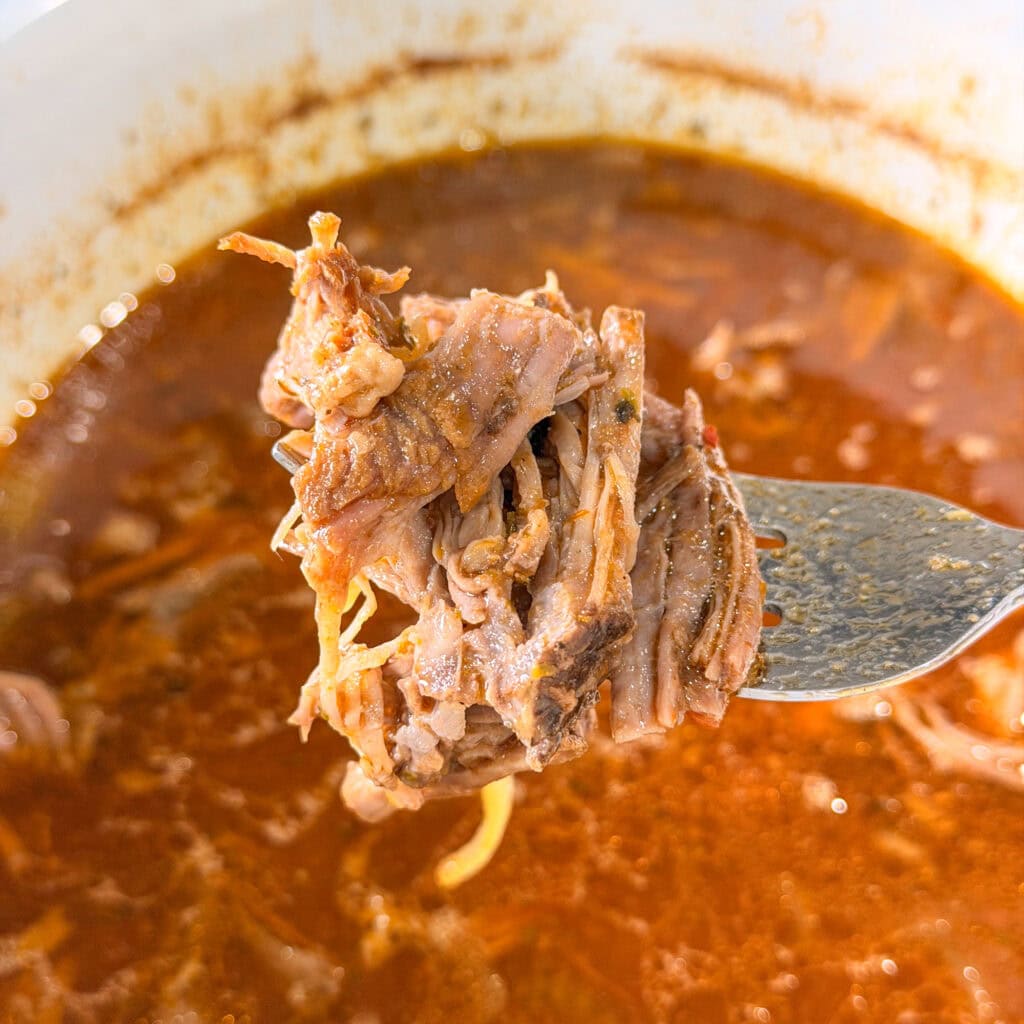 A fork holds a serving of shredded beef above a bowl of rich, reddish-brown broth, reminiscent of Crock Pot Italian Beef Sandwiches. The meat is tender and juicy, with visible seasonings and strands.