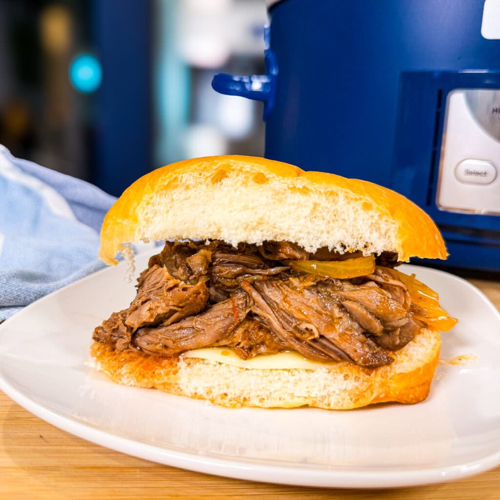 A Crock Pot Italian Beef Sandwich sits on a white plate, piled with shredded beef, cheese, and pickles on a bun. In the background, a blue appliance and a blue-and-white cloth are visible.