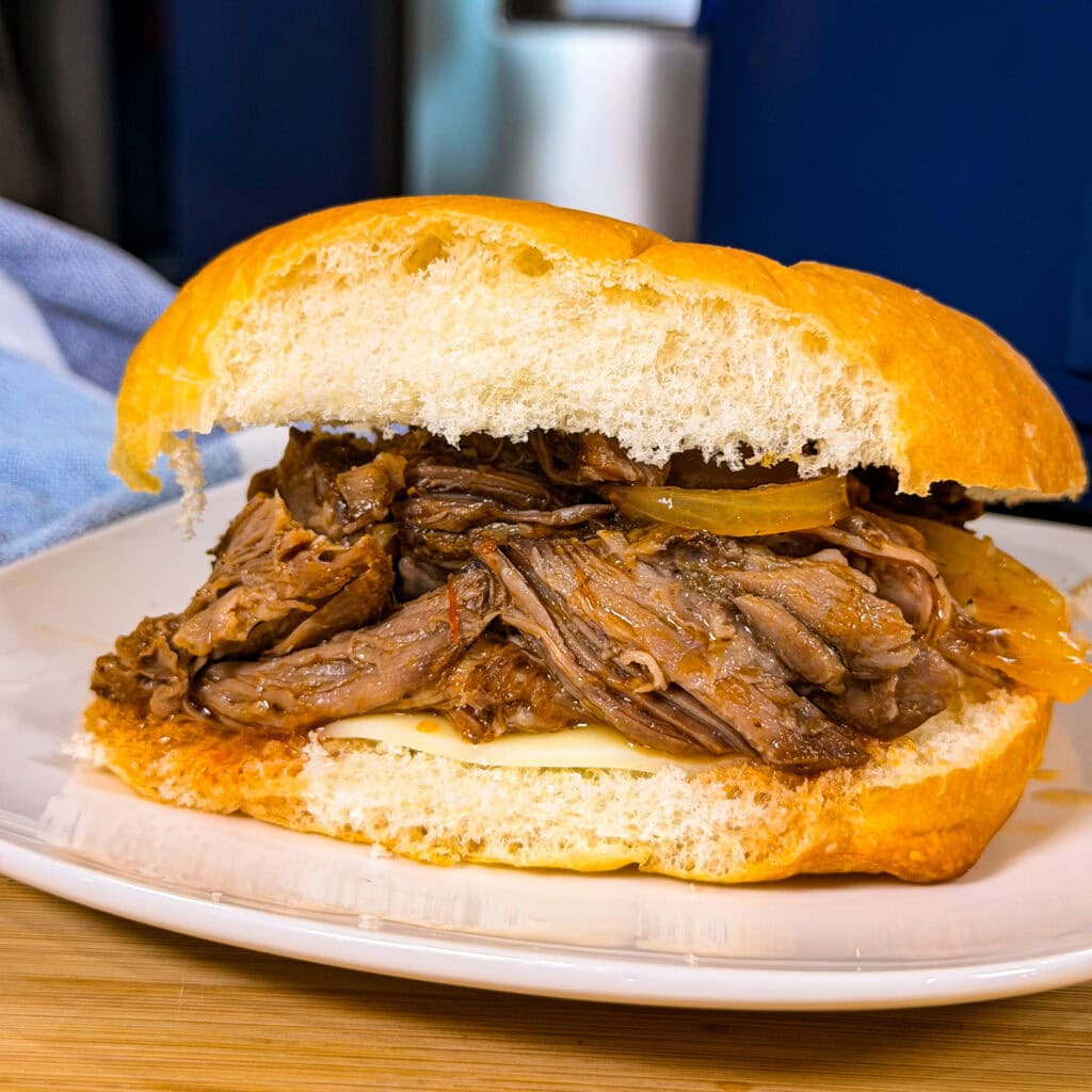 A close-up of Crock Pot Italian Beef Sandwiches filled with shredded beef and onions on a round bun, served on a white plate.