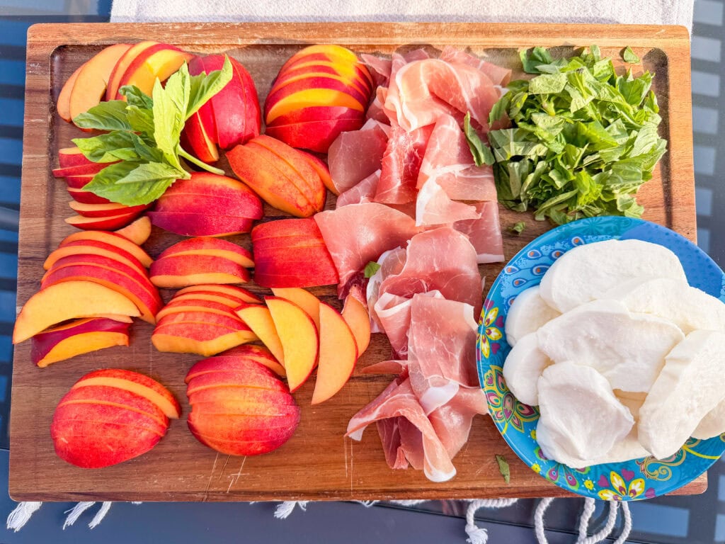 A wooden board with sliced peaches, fresh basil and mint, prosciutto, and a plate of sliced mozzarella cheese—perfect for serving as a Nectarine Prosciutto Caprese Salad.