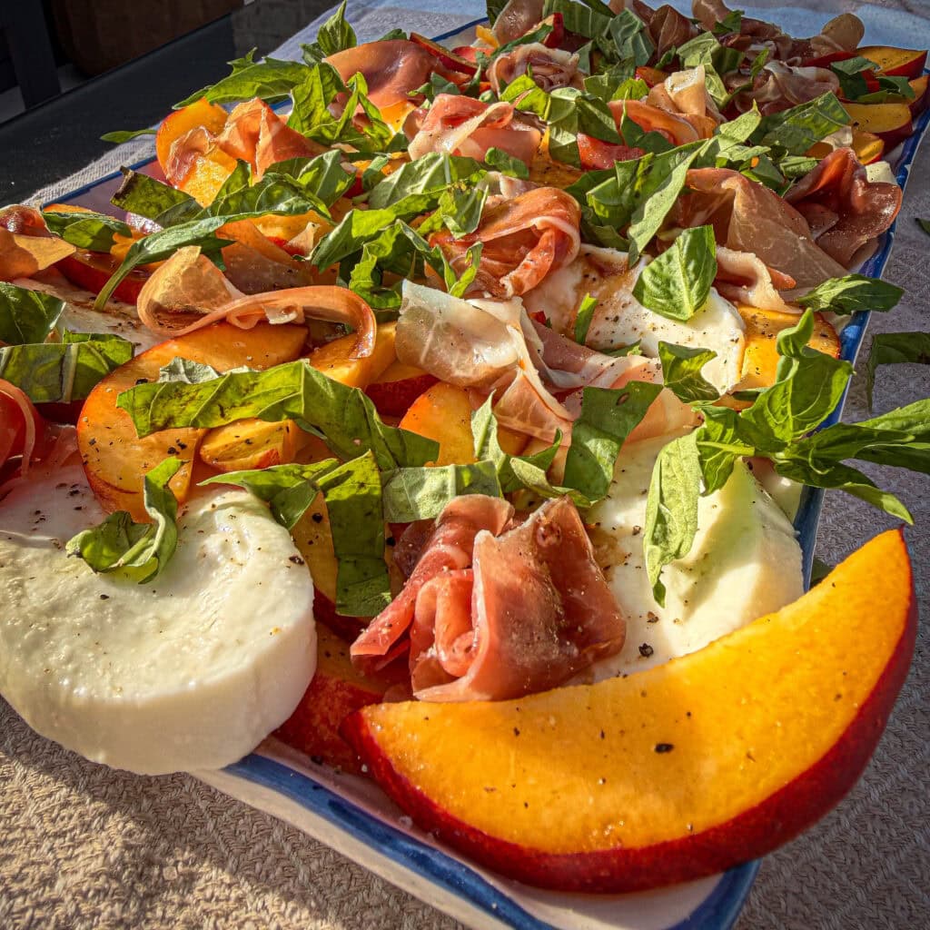 A close-up of a vibrant Nectarine Prosciutto Caprese Salad with fresh mozzarella slices, prosciutto, nectarine, tomato, and basil, drizzled with olive oil and black pepper on a rectangular blue dish.