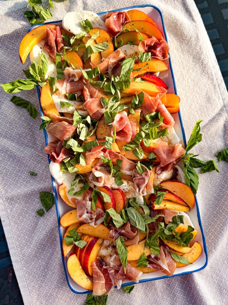 A rectangular dish filled with Nectarine Prosciutto Caprese Salad—sliced peaches, fresh mozzarella, prosciutto, and chopped basil—rests on a light-colored textured cloth in bright sunlight.