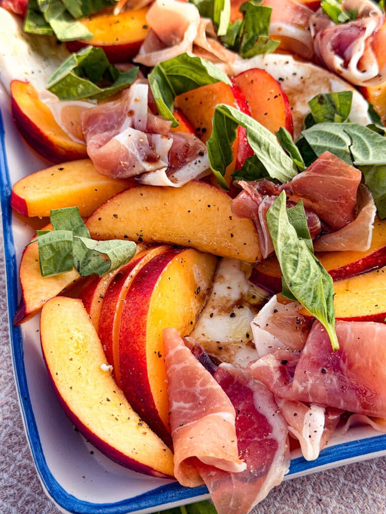 A close-up of a Nectarine Prosciutto Caprese Salad features sliced peaches, prosciutto, fresh basil leaves, and mozzarella, all seasoned with black pepper and arranged in a blue-edged dish.