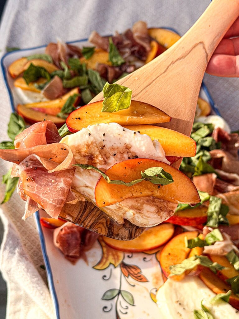 A wooden spatula holds a serving of Nectarine Prosciutto Caprese Salad with fresh mozzarella, peach slices, prosciutto, basil, and balsamic over a decorative platter with more salad in the background.