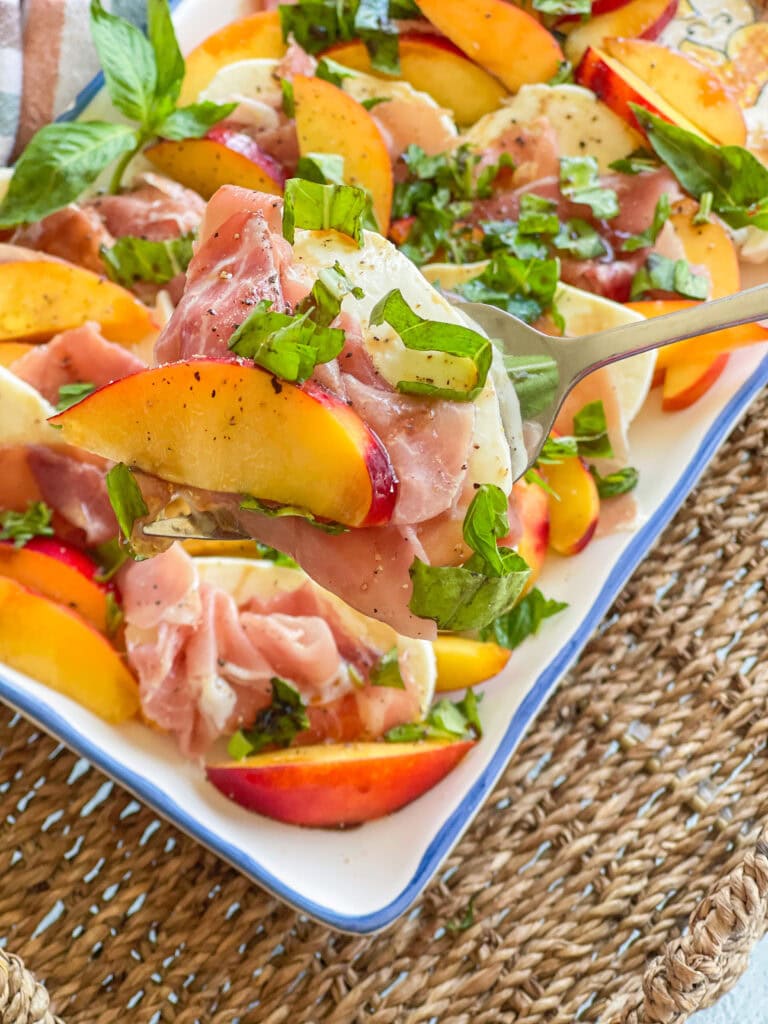 Plated nectarine Caprese salad with slices of fresh mozzarella and prosciutto, garnished with julienned basil and drizzled with olive oil and balsamic glaze, arranged on a white serving platter.