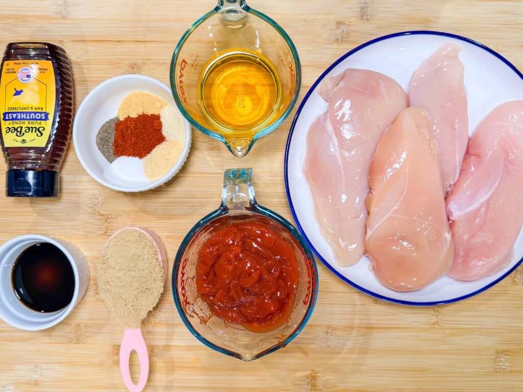 Raw chicken breasts on a plate surrounded by honey, seasonings, brown sugar, ketchup, soy sauce, and oil in small bowls—everything you need to make flavorful Crock Pot BBQ Chicken on a wooden surface.