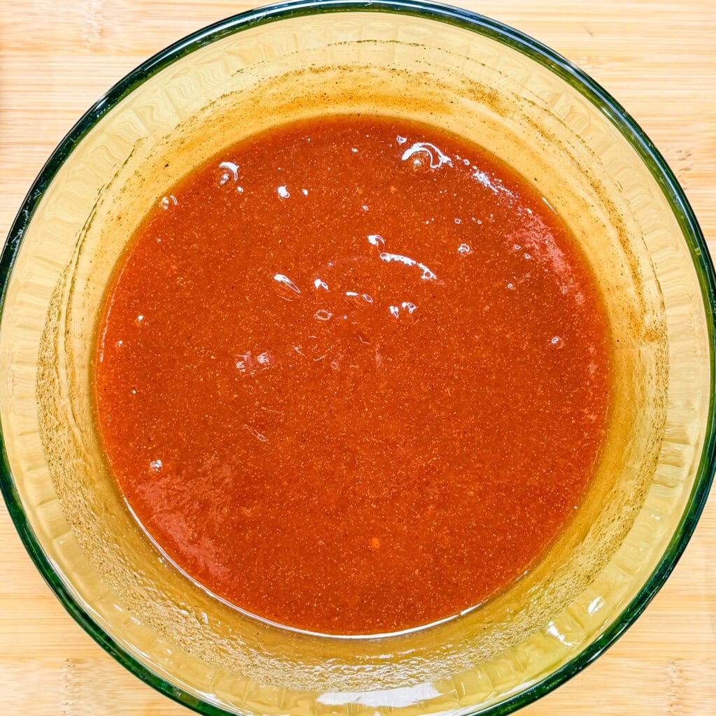 A clear glass bowl filled with a smooth, reddish-brown sauce or dressing—perfect for Crock Pot BBQ Chicken—sits on a light wooden surface. The mixture has a shiny, slightly thick consistency, with some small bubbles and specks visible.