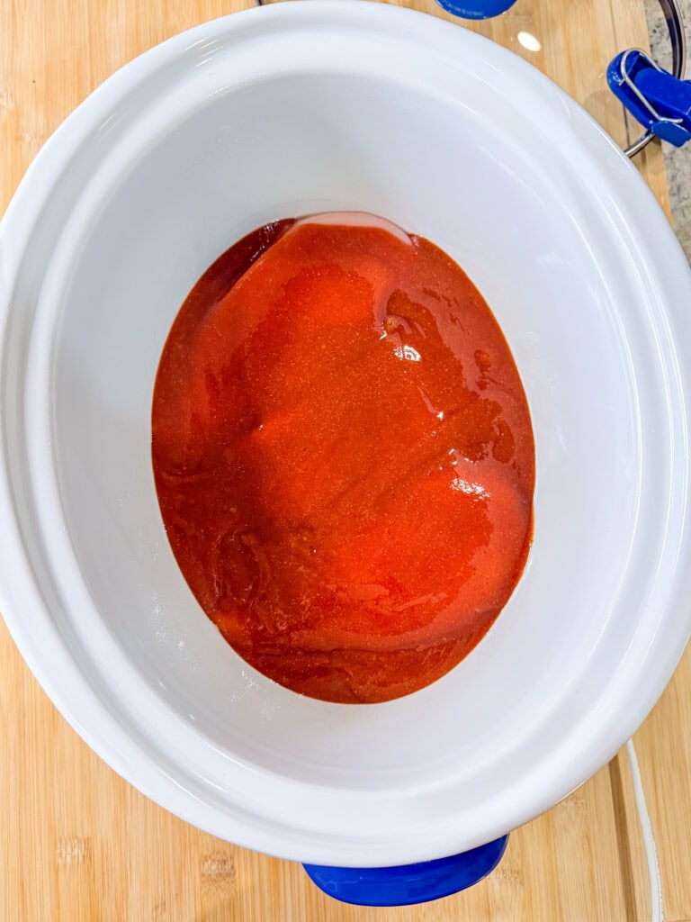 A white Crock Pot sits on a wooden surface, filled with a layer of red sauce spread across the bottom—perfect for starting a delicious BBQ Chicken recipe.