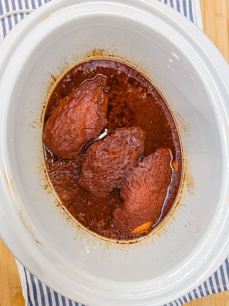 Three pieces of beef in a rich, dark red sauce simmer in a white slow cooker, placed on a blue and white striped cloth—perfect inspiration for your next Crock Pot BBQ Chicken dish.