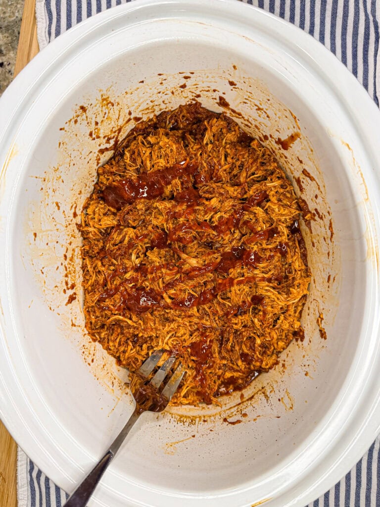 Pulled Crock Pot BBQ Chicken with sauce sits in a white slow cooker, a metal fork resting inside. The cooker is placed on a blue-and-white striped cloth.