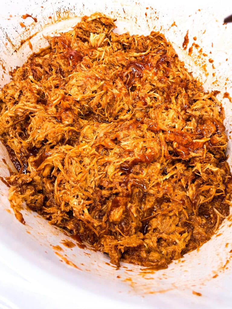 Shredded barbecue pulled pork mixed with sauce in a white slow cooker—just like preparing Crock Pot BBQ Chicken, but with tender pork for a savory twist.