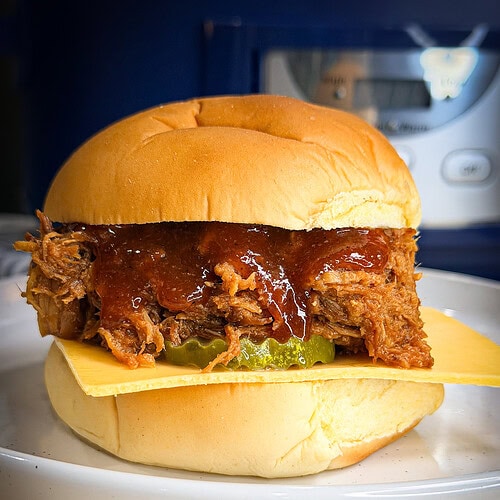 A delicious sandwich featuring Crock Pot BBQ Chicken or pulled pork, topped with barbecue sauce, pickles, and a slice of cheese on a soft bun, served on a white plate.