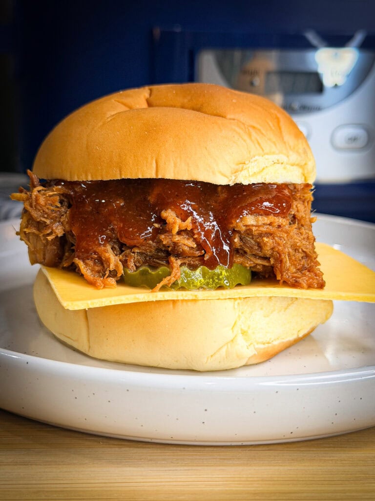 A delicious sandwich featuring Crock Pot BBQ Chicken or pulled pork, topped with barbecue sauce, pickles, and a slice of cheese on a soft bun, served on a white plate.