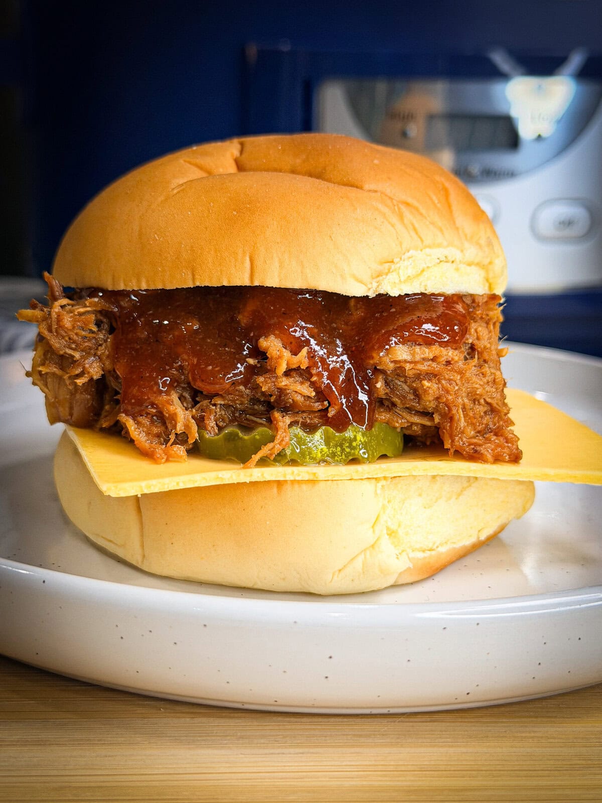 A delicious sandwich featuring Crock Pot BBQ Chicken or pulled pork, topped with barbecue sauce, pickles, and a slice of cheese on a soft bun, served on a white plate.