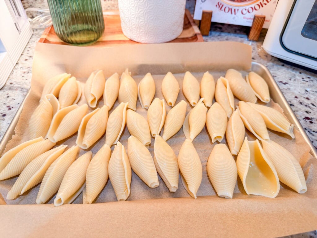 Uncooked jumbo pasta shells are arranged in rows on a parchment-lined baking sheet, ready to become delicious Chicken Alfredo Stuffed Shells. The tray rests on a speckled countertop with kitchen containers and cookbooks in the background.