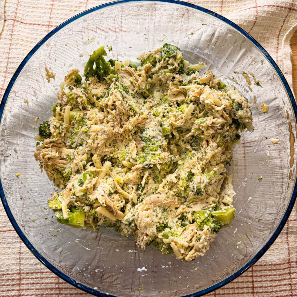 A glass bowl filled with a mixture reminiscent of Chicken Alfredo Stuffed Shells—shredded chicken, broccoli, cheese, and seasonings—sits on a beige checkered cloth.