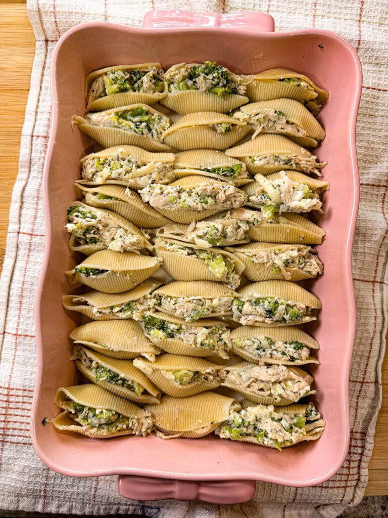 Unbaked pasta shells stuffed with a creamy ricotta and spinach mixture, reminiscent of comforting Chicken Alfredo Stuffed Shells, are arranged neatly in rows inside a pink baking dish, which sits on a checkered cloth.