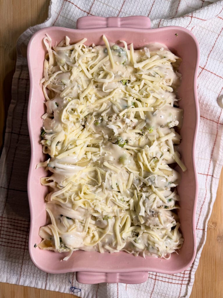 A pink baking dish filled with unbaked Chicken Alfredo Stuffed Shells, topped with shredded cheese and creamy sauce, sits on a checkered towel on a wooden surface.