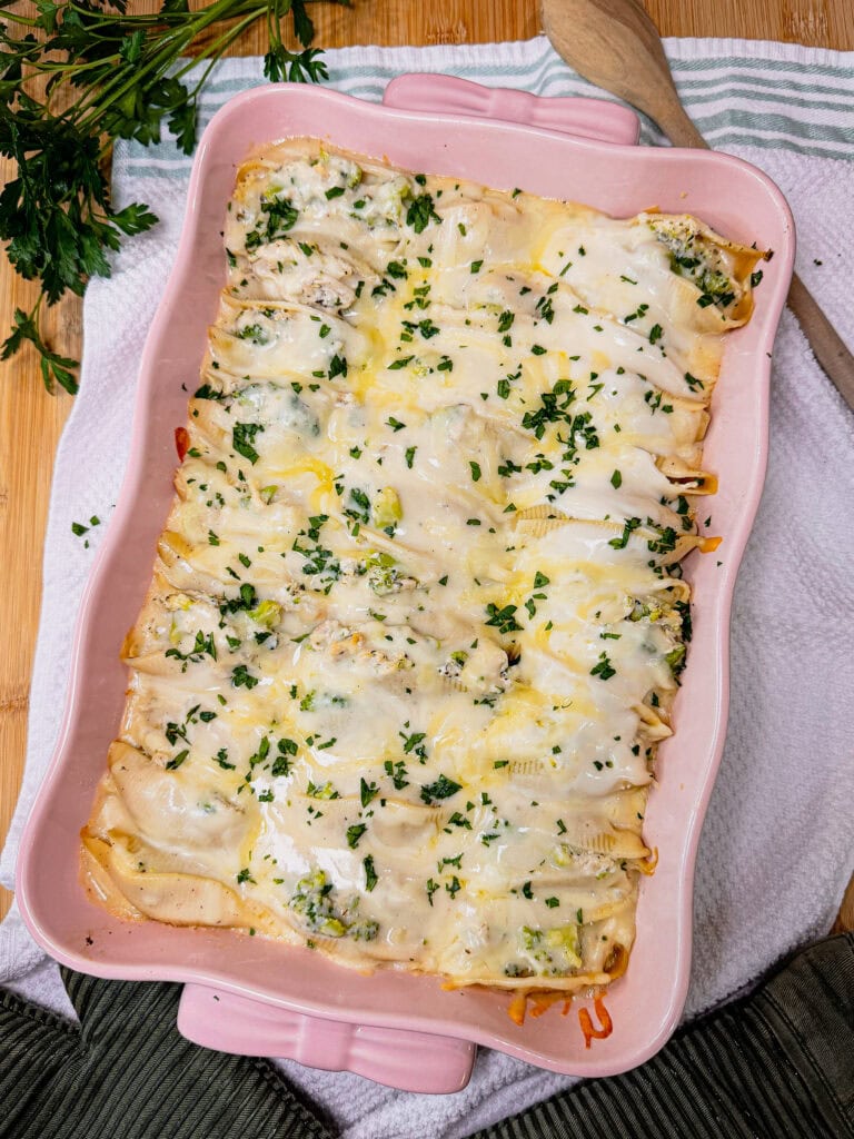 A pink baking dish filled with Chicken Alfredo Stuffed Shells, covered in melted cheese and sprinkled with chopped herbs, sits on a cloth, with a wooden spoon and parsley nearby.
