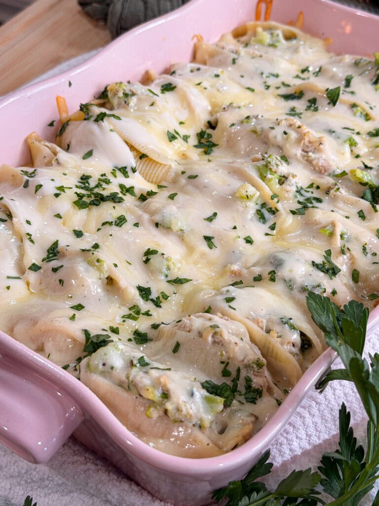 Chicken Alfredo Stuffed Shells in a pink baking dish, topped with melted cheese and chopped fresh parsley. The creamy filling is flecked with herbs and likely includes tender chicken along with spinach or zucchini. This comforting dish rests on a white cloth, ready to serve.